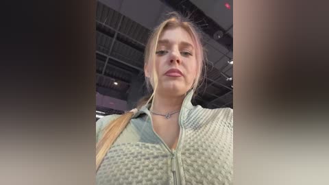 Video of a fair-skinned blonde woman with a slight smile, wearing a light blue, textured jacket, indoors with exposed beams and industrial lighting.