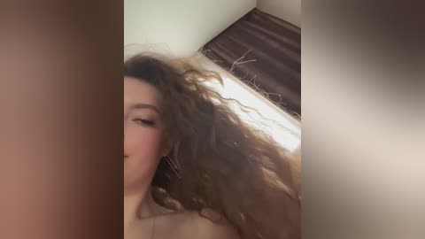 A video of a young woman with long, wavy brown hair, wearing large hoop earrings, captured from a low angle, emphasizing her head and shoulders. The background shows a light-colored wall and a partially visible wooden shelf.