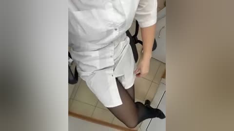 A video of a person in a white dress, black tights, and black shoes, seated in a beige-tiled room, holding a white clipboard.