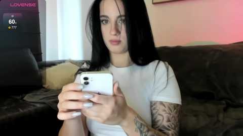 Video of a young woman with long black hair, wearing a white t-shirt, holding a phone, showing her tattooed arm, sitting on a dark couch in a dimly lit room.