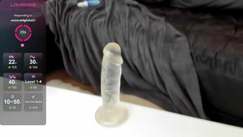 Video of a clear, translucent dildo with a bulbous head and textured shaft, lying on a black, crumpled fabric surface. The background includes a digital screen displaying a virtual thermometer with a temperature of 30\u00b0C.