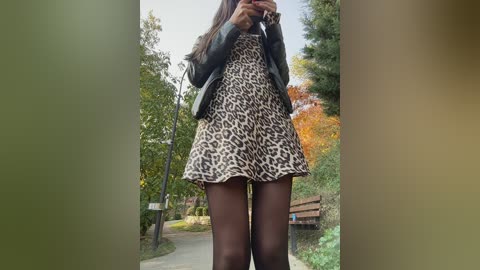 Video of a young woman with long brown hair, wearing a leopard print dress, black tights, and a denim jacket, standing outdoors in a park with trees and a wooden bench in the background.