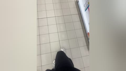 Video of a person's feet walking on a tiled floor in a restroom. The person is wearing black pants and white sneakers. A white trash can with colorful stickers is visible in the background.