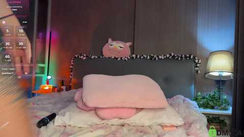 Video of a plush pink cat on a bed with white pillows, surrounded by vibrant LED lights and a green potted plant, in a modern, cozy bedroom.