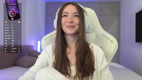 Video of a young, fair-skinned woman with long brown hair, wearing white headphones, seated on a white gaming chair in a modern, dimly lit bedroom.