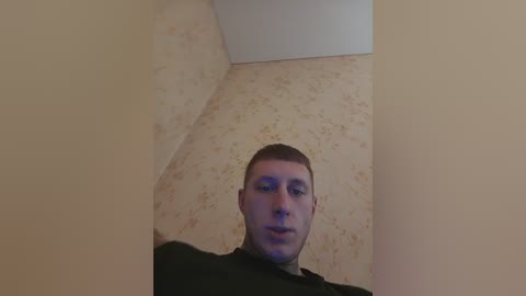 Video of a young man with short hair, fair skin, and a neutral expression, standing in a corner of a room with beige wallpaper and textured floral pattern.