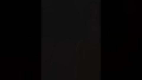 A dim, grainy video of a tall, slender figure in a black jacket, partially obscured by shadows, standing against a dark background, possibly in a nightclub.