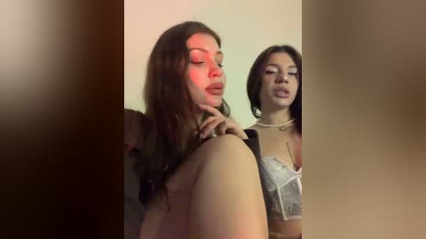 Video of two women, one topless, the other in lingerie, in a dimly lit room, with a red light casting a sultry glow.