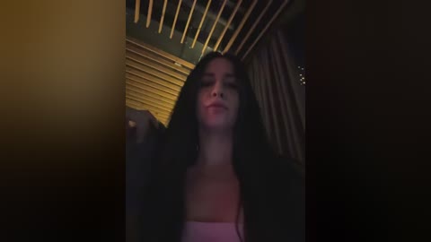 Video of a young woman with long dark hair, wearing a pink top, standing in a dimly lit room with wooden slatted ceiling and drapes.