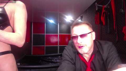 Video of a man with short hair, wearing sunglasses and a black jacket, sitting on a black leather couch in a dimly lit room with red and black walls and BDSM gear hanging on the wall.