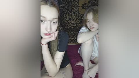 Video of a young woman with fair skin and shoulder-length blonde hair, wearing a black top, and a girl with similar features, sitting on a red bedspread, against a patterned wall.