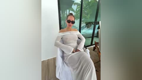 Video of a fair-skinned woman with light brown hair, wearing a white off-shoulder dress, sitting on a chair, with sunglasses, in a modern room with large windows and wooden flooring.