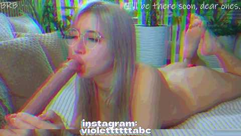 A vibrant, colorful video of a nude, fair-skinned woman with long blonde hair, wearing glasses, lying on a couch. She is licking an erect penis. Background shows potted plants and text overlays.