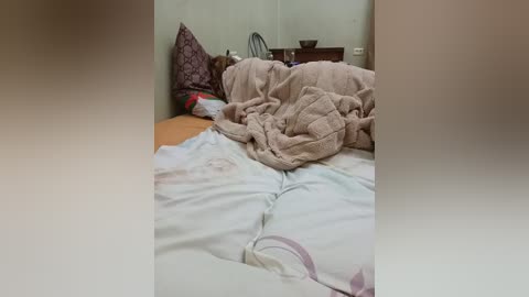 A video of a messy bedroom with unmade bed covered in beige and white sheets, pillows scattered, and a wooden nightstand with a bowl and small plant in the background.