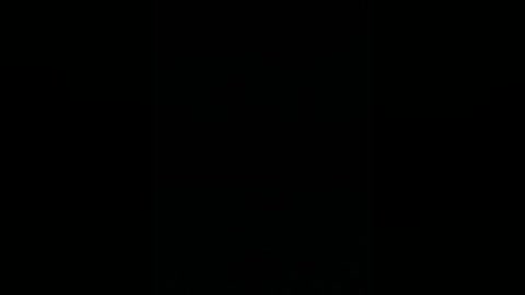 A minimalist, black-and-white video of a lone, dark-haired woman with a serious expression, standing against a stark black background. The image emphasizes simplicity and solitude.