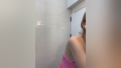 A video of a woman in a bathroom, seen from the side, with a pink towel wrapped around her shoulders. She has fair skin and dark hair. White tiled walls and a closed door are visible in the background.
