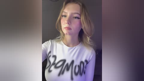 Video of a young Caucasian woman with light skin, shoulder-length blonde hair, and a slight smile. She wears a white T-shirt with \"Semora\" printed in black letters. Background is dark and blurry.