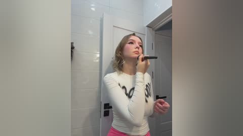 Video of a young, light-skinned woman with shoulder-length blonde hair, wearing a white long-sleeved shirt with black writing and pink pants, applying makeup in a white-tiled bathroom.