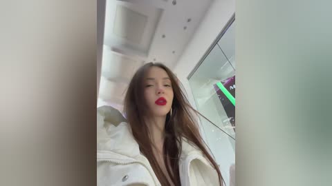 A video of a young woman with long brown hair, red lipstick, and white robe, standing in a modern, white-tiled bathroom with glass doors and a green neon sign in the background.