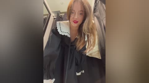 A video of a young woman with light skin, long blonde hair, and bright red lipstick, wearing a black graduation gown with a white collar, standing in a hallway.