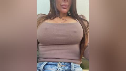 A video of a woman with long brown hair, wearing a tight, beige tank top that accentuates her large breasts and high-waisted, light blue jeans. The background is blurred, focusing on her upper body.