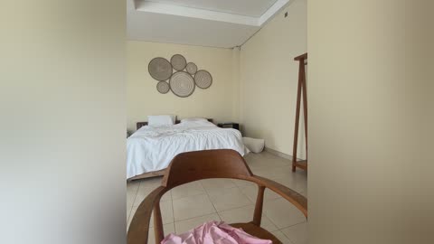 Video of a minimalist, beige-walled bedroom with a white bed, wooden chair, and decorative wall art.