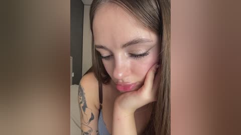 Video of a young Caucasian woman with straight brown hair, wearing minimal makeup, resting her head on her hand. She has a tattoo on her left arm. Background features a plain wall and a glimpse of a bed.