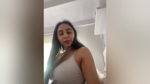 A video of a young Latina woman with long, dark hair, wearing a beige spaghetti-strap tank top, standing in a modern, bright, white room with minimalist decor.