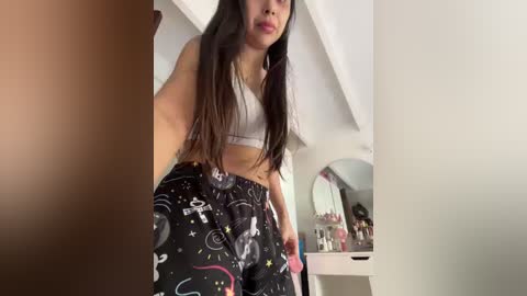 Video of a young woman with long black hair, wearing a white sports bra and black leggings with cosmic designs, standing in a brightly lit bedroom with a vanity and mirror in the background.