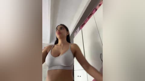 A video captures a young woman with medium skin tone and long black hair, wearing a white sports bra, standing in a narrow hallway. The background features white walls and a partially visible door with pink trim.