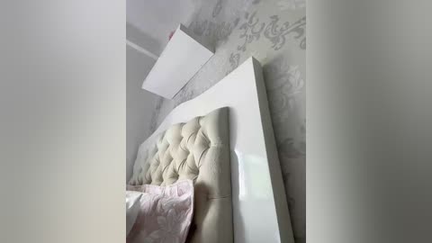A video of a modern, minimalist bedroom with a plush, light beige tufted headboard, white walls, and a subtle gray floral wallpaper. The bed is made with pink and white bedding, and a white ceiling fan is visible.