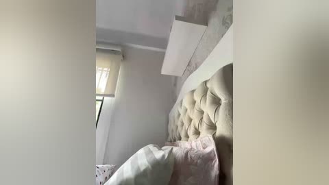 Video of a minimalist bedroom with a beige tufted headboard, white walls, and a small window with a beige curtain. The room is softly lit, creating a serene ambiance.