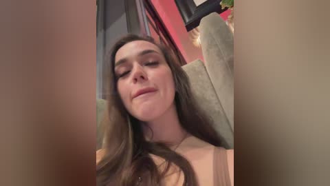 Video of a smiling, fair-skinned woman with long brown hair, wearing a beige top, sitting in a plush green chair with a red and black background.