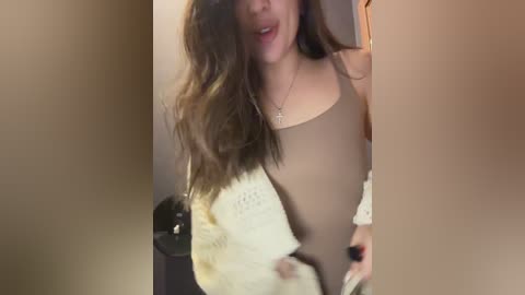 Video of a young woman with long, wavy brown hair, wearing a beige sleeveless top, standing indoors, with blurred background.