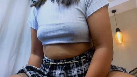 Video of a woman with medium brown skin, wearing a tight, light blue crop top revealing a small waist and a plaid skirt. She sits indoors with a pendant light hanging in the background.
