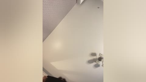 Video of a ceiling fan in a white room, taken from below. The fan has three blades and is mounted on a textured white ceiling. The image is slightly blurred, giving a sense of motion.