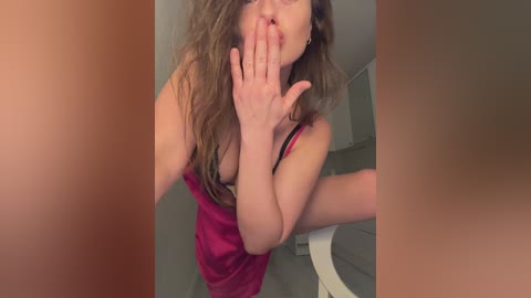 A video of a young woman with long, wavy brown hair, wearing a pink camisole, covering her mouth, and touching her nose. She's in a bathroom with a white toilet and grey tiled floor.