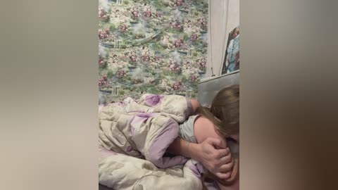 Video of a young girl with light skin, brown hair, wearing a white shirt, crying and hugging a pink and white blanket in a bedroom with floral wallpaper.