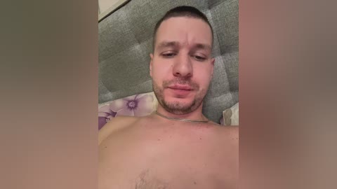 Video of a shirtless, muscular man with short brown hair and a beard, wearing a silver necklace, lying on a bed with a floral-patterned purple pillow.