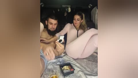 Video of a bearded man with a beard, shirtless, and a woman in a pink jumpsuit, both sitting in a car, holding a camera, with a box of donuts nearby.