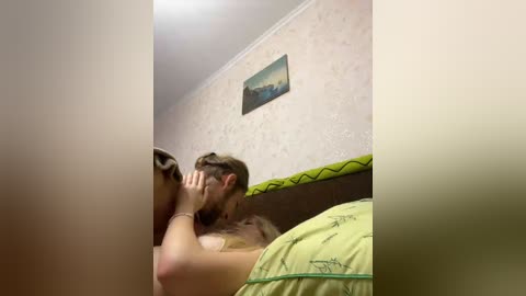 Video of a man with light brown hair, wearing a green shirt, kissing a woman with light brown hair, both sitting on a bed with patterned sheets.