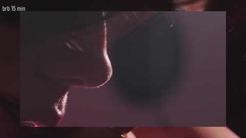 A close-up video of a person's face, focusing on the nose and mouth, with a blurred background. The image has a dim, reddish hue, emphasizing the shadowy, intimate atmosphere.