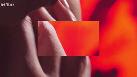 A close-up video of a person's hand holding a rectangular, bright orange object against a blurred, fiery red background. The hand is positioned on the left side, fingers gently gripping the object. The image is labeled \"bri 15 min\" in the top left corner.