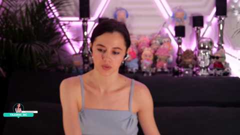 Video of a young woman with fair skin and dark hair, wearing a light blue sleeveless top, standing in front of a colorful, decorated stage with balloons and floral arrangements.