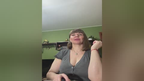 Video of a middle-aged woman with shoulder-length brown hair, wearing glasses and a gray V-neck top, sitting indoors with green walls, a shelf, and a plant in the background.