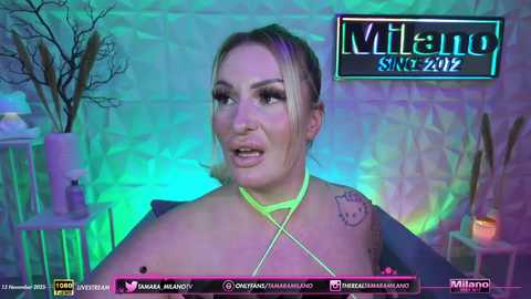 Video of a blonde woman with a medium build, wearing a neon yellow halter top, standing in a dimly lit room with green and purple lighting, displaying tattoos on her shoulders and collarbone.