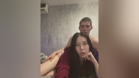 Video of an Asian woman with long black hair, wearing a red shirt, being held by a white man with short brown hair, in a dimly lit room with a textured gray wall and a visible air conditioner.