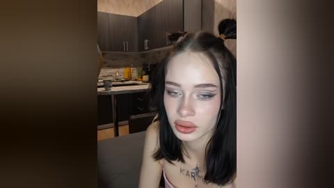 Video of a pale-skinned, black-haired woman with two pigtails, wearing a spaghetti-strap top, standing in a dimly lit kitchen with dark cabinets, a stove, and a countertop.