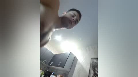 Video of a naked man with short dark hair, standing in a dimly lit room with a ceiling light, dark cabinets, and a blurred background.