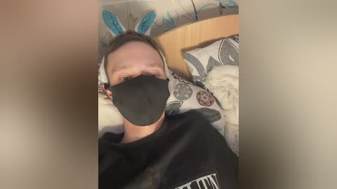 A video of a young man lying in bed, wearing a black face mask, blue bunny ears, and a black t-shirt, surrounded by white and patterned bedding.